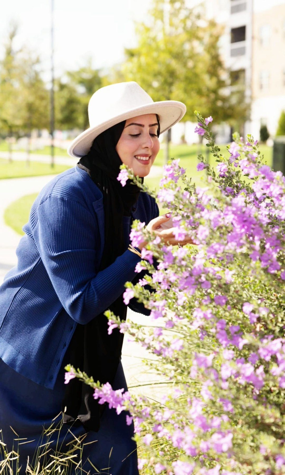 Huda in a cream hat and navy cardigan, eyes closed and smiling, leaning into a flowering lavender bush