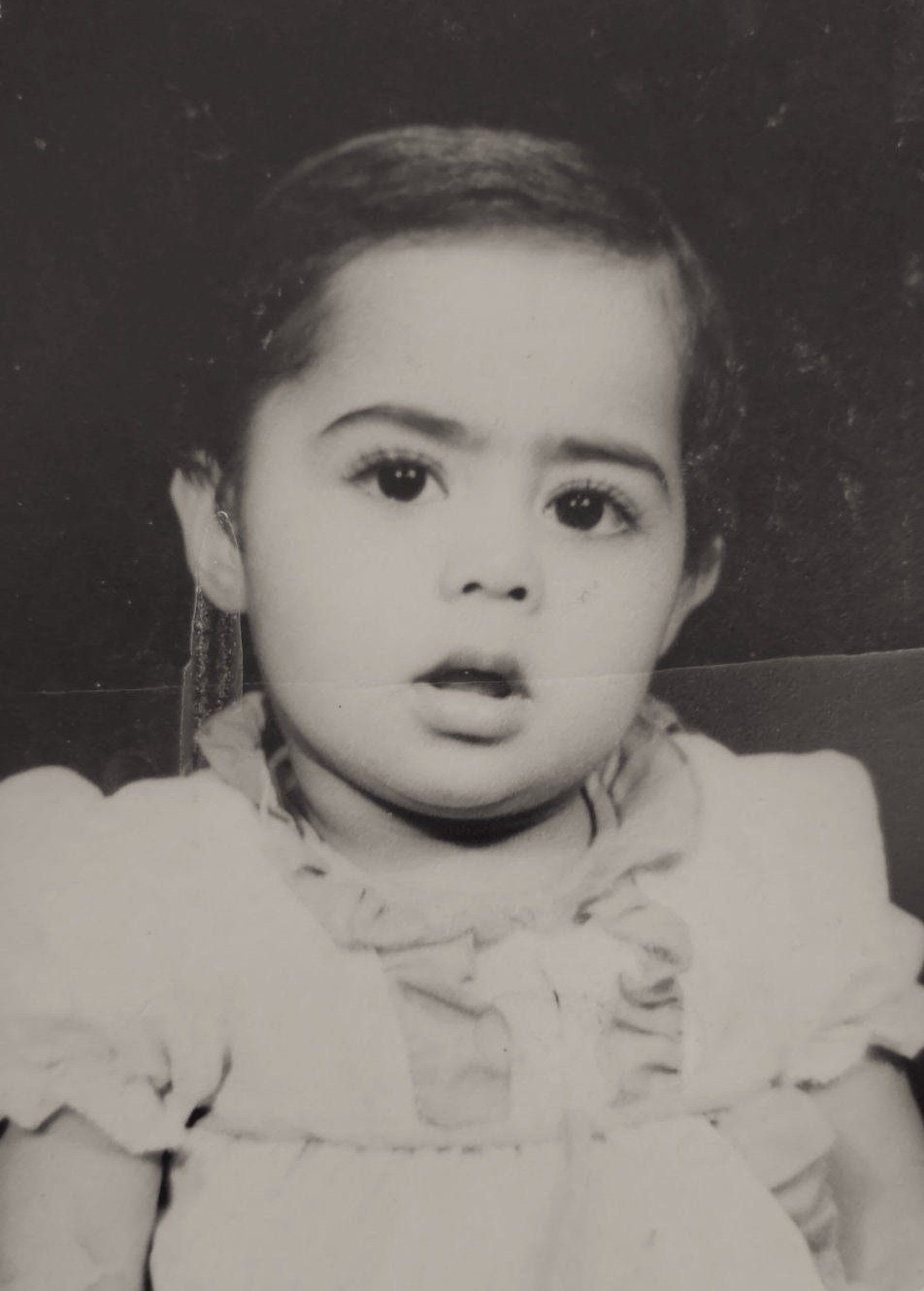 Black-and-white childhood photograph of Huda as a toddler, wide-eyed, in a ruffled white dress