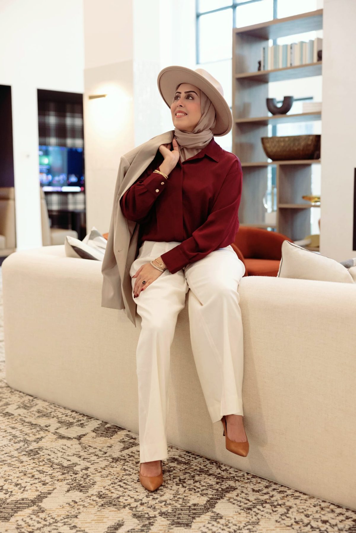 Huda Mooswi in a burgundy blouse and cream trousers, jacket over her shoulder, looking upward with a smile