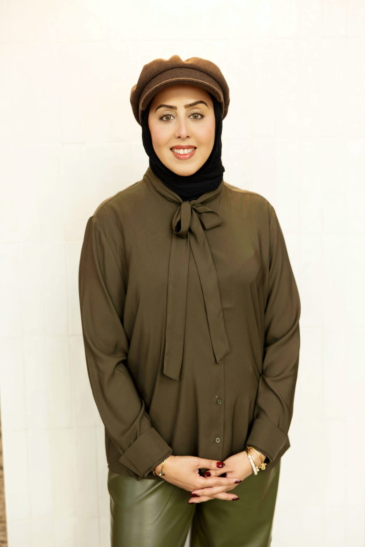Huda Mooswi standing in an olive blouse and brown cap, hands clasped, smiling at the camera